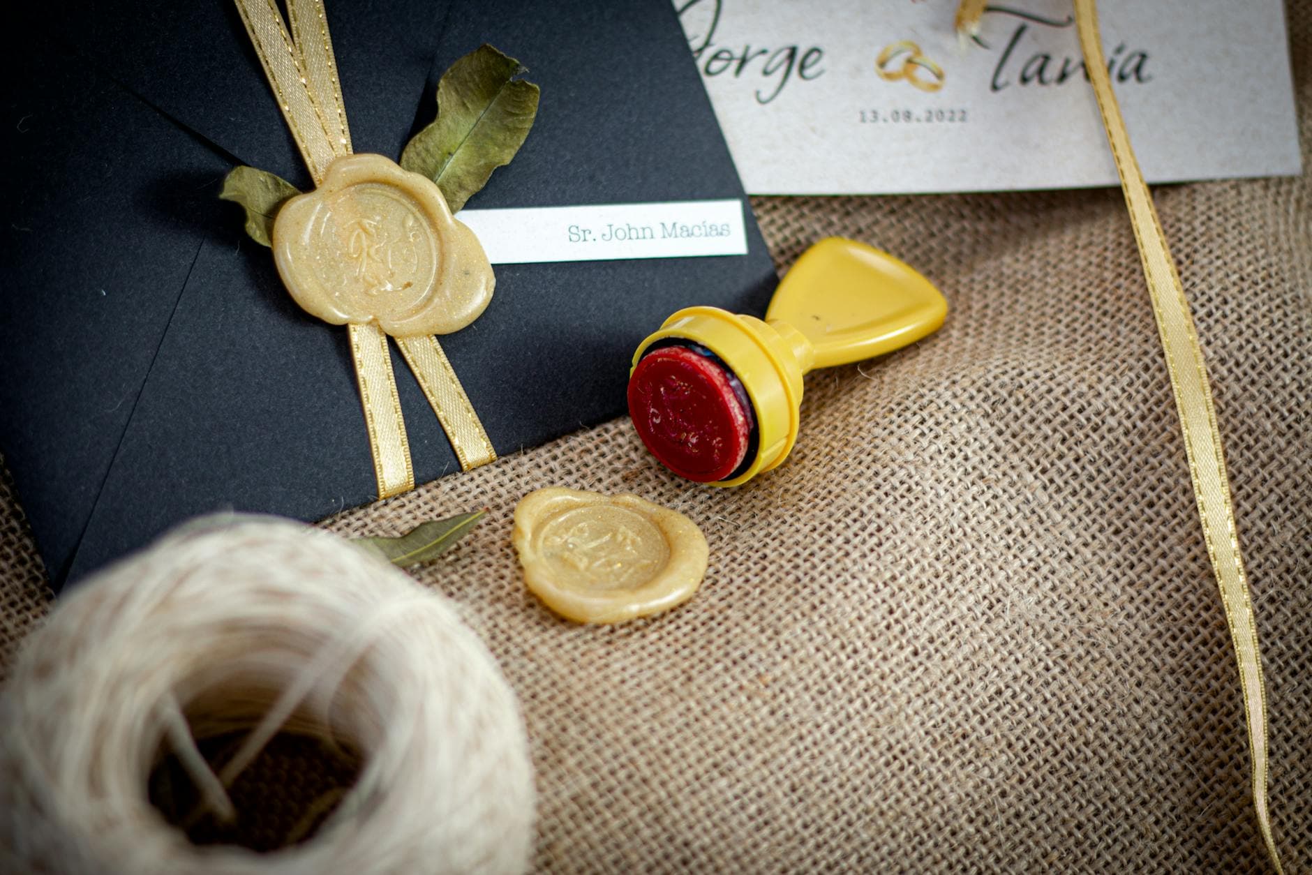 Close-up of a black envelope with a decorative wax seal, ribbon, and stamp on burlap.