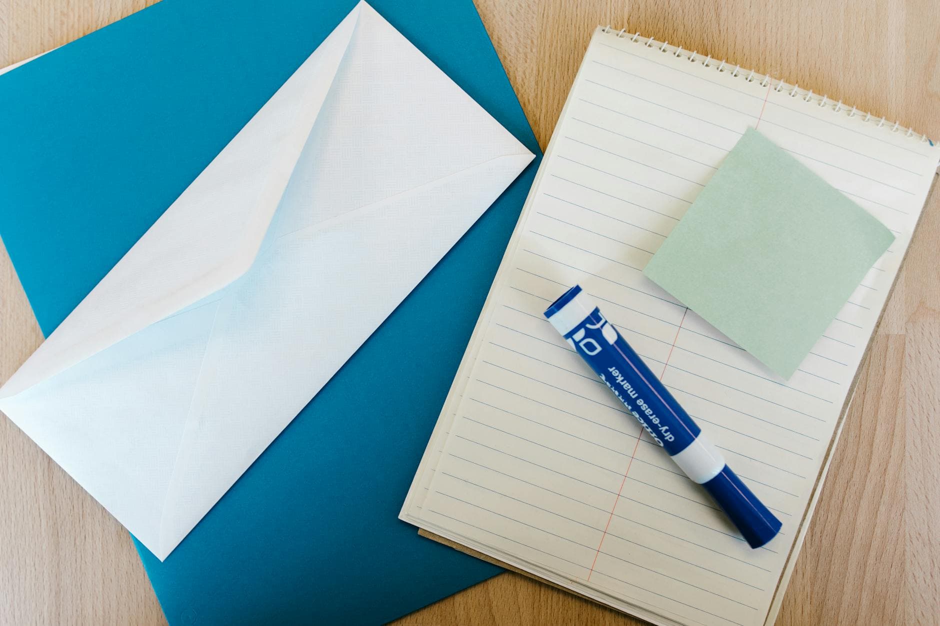 Flat lay of office supplies on a wooden desk, including a notepad, envelope, marker, and sticky note.